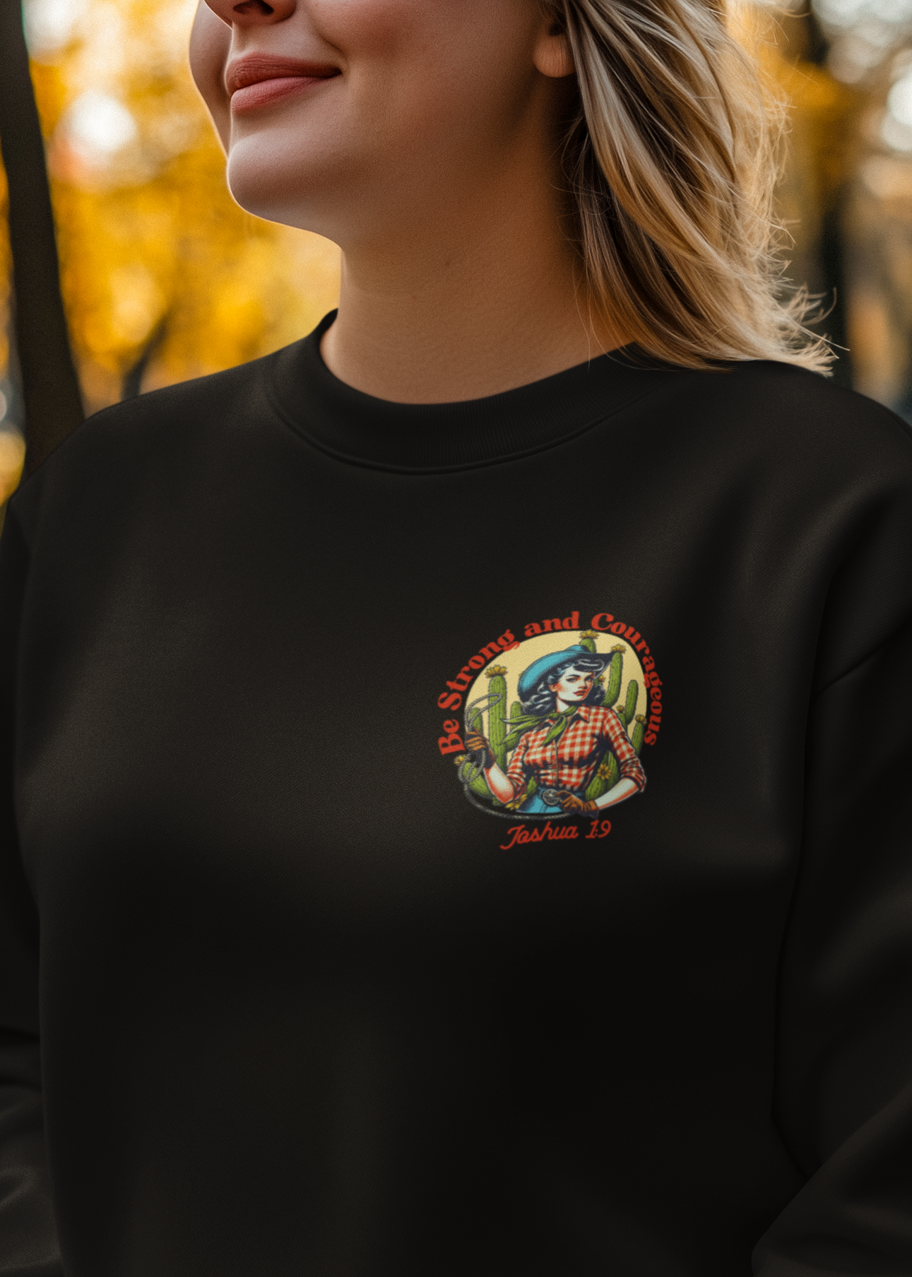 A woman wearing a black crewneck sweatshirt with a colorful graphic on the left chest, photographed outdoors in a park with autumn trees and fallen leaves.