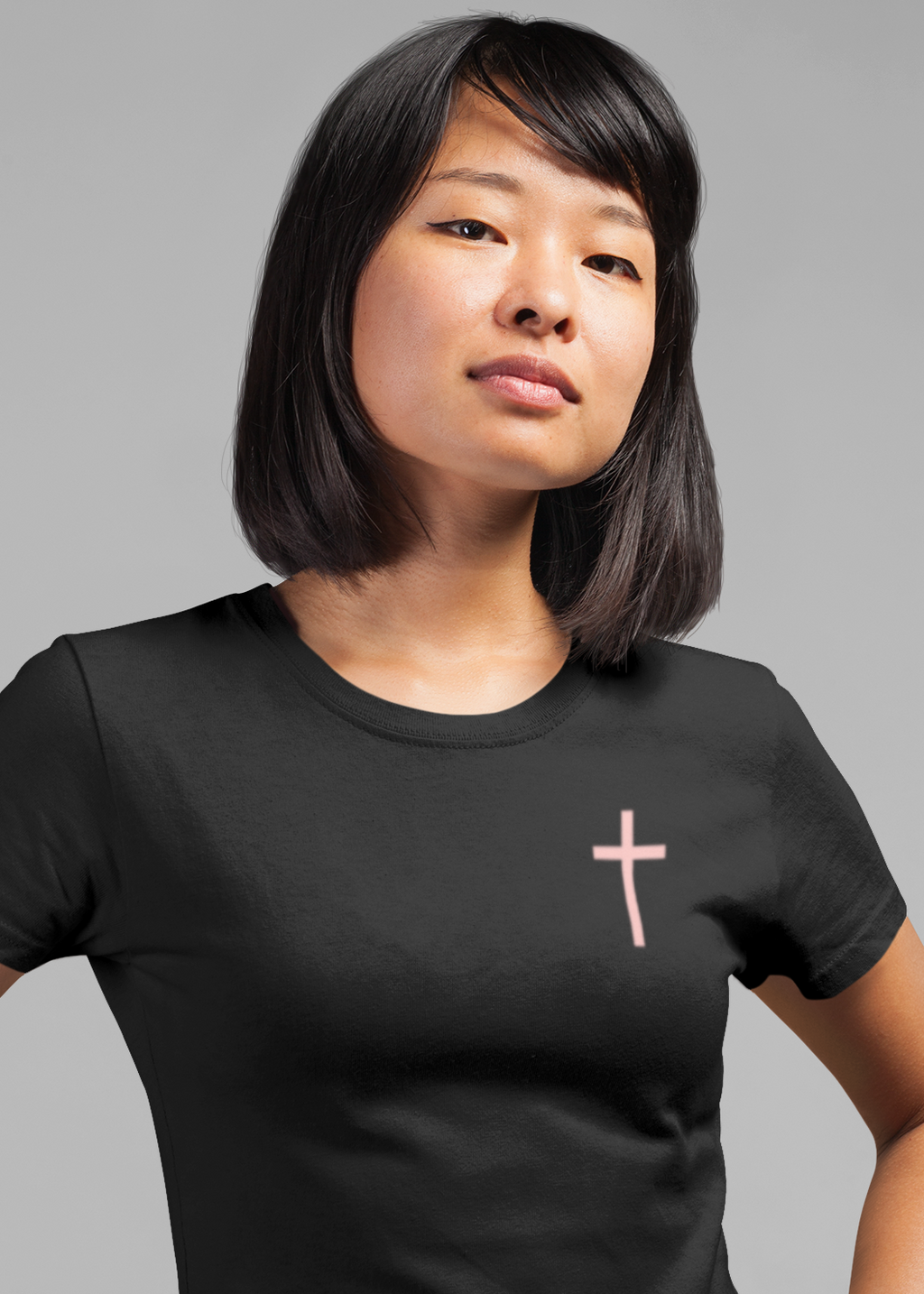 A woman with a straight dark bob haircut wearing a black crewneck t-shirt with a small light pink cross on the left chest, posing with hands on hips against a solid gray background.