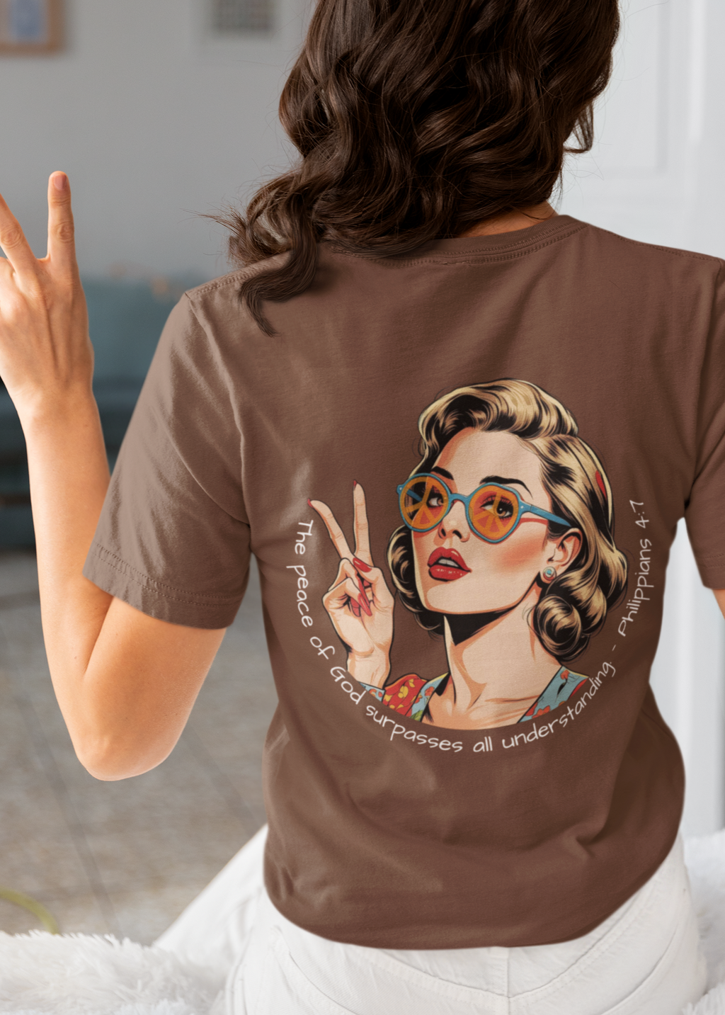 Person wearing a brown t-shirt with a graphic design of a woman with glasses and text, making peace signs. White Christian graphic tee with retro 70s inspired woman holding up a peace sign and wearing peace sign glasses. Inspired by the scripture Philippians 4:7. 