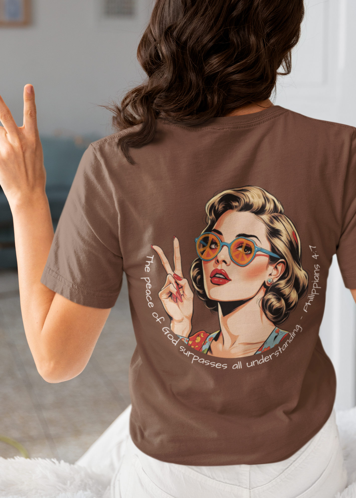 Person wearing a brown t-shirt with a graphic design of a woman with glasses and text, making peace signs. White Christian graphic tee with retro 70s inspired woman holding up a peace sign and wearing peace sign glasses. Inspired by the scripture Philippians 4:7. 