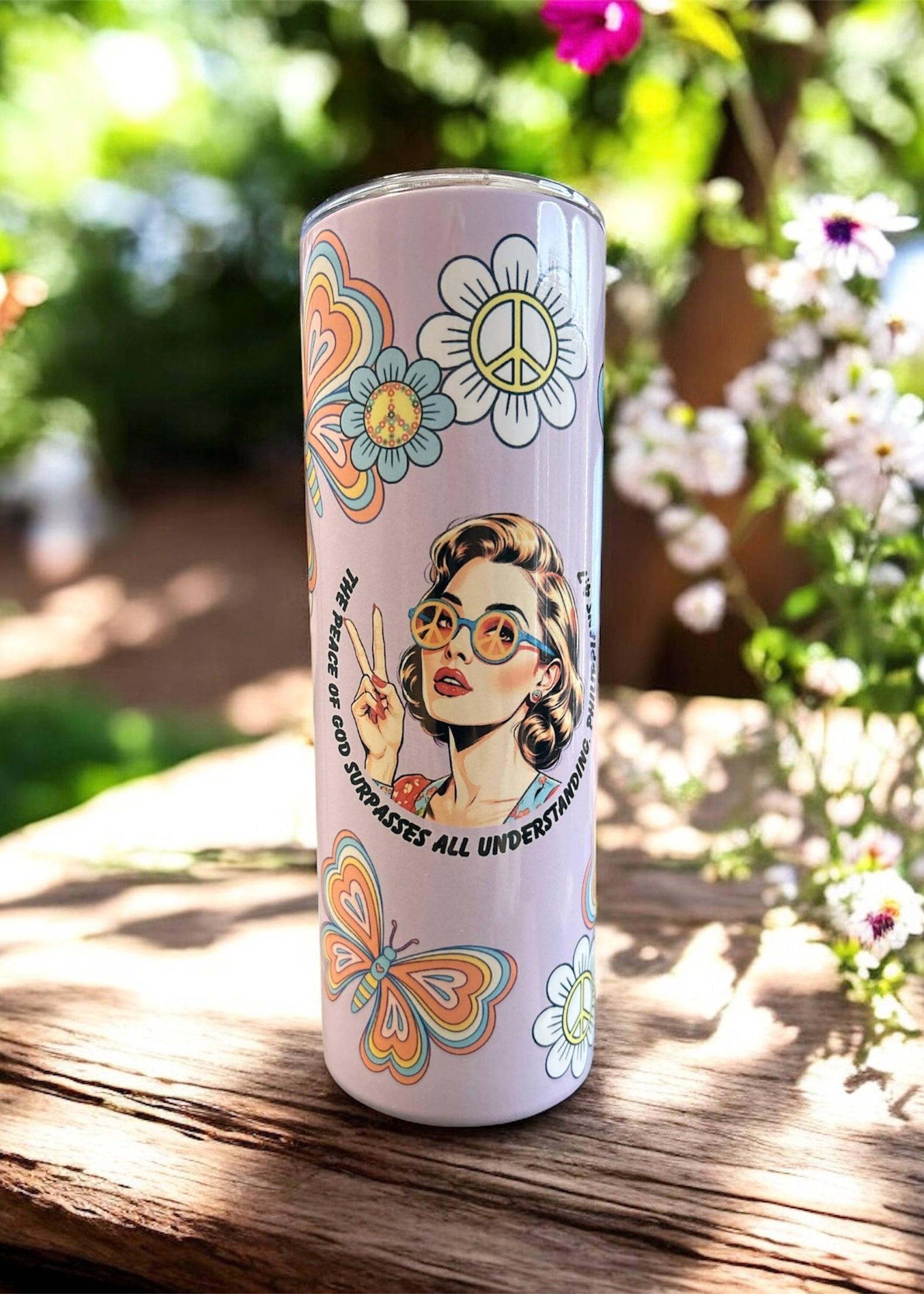 Tumbler with artistic design featuring a woman, flowers, and peace symbol on a wooden surface with a natural background.