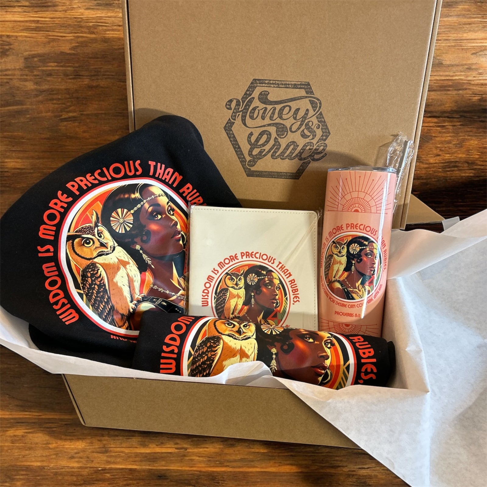 Box with 'Honey's Grace' branding containing a black t-shirt, a white card, and a pink product on a wooden surface.