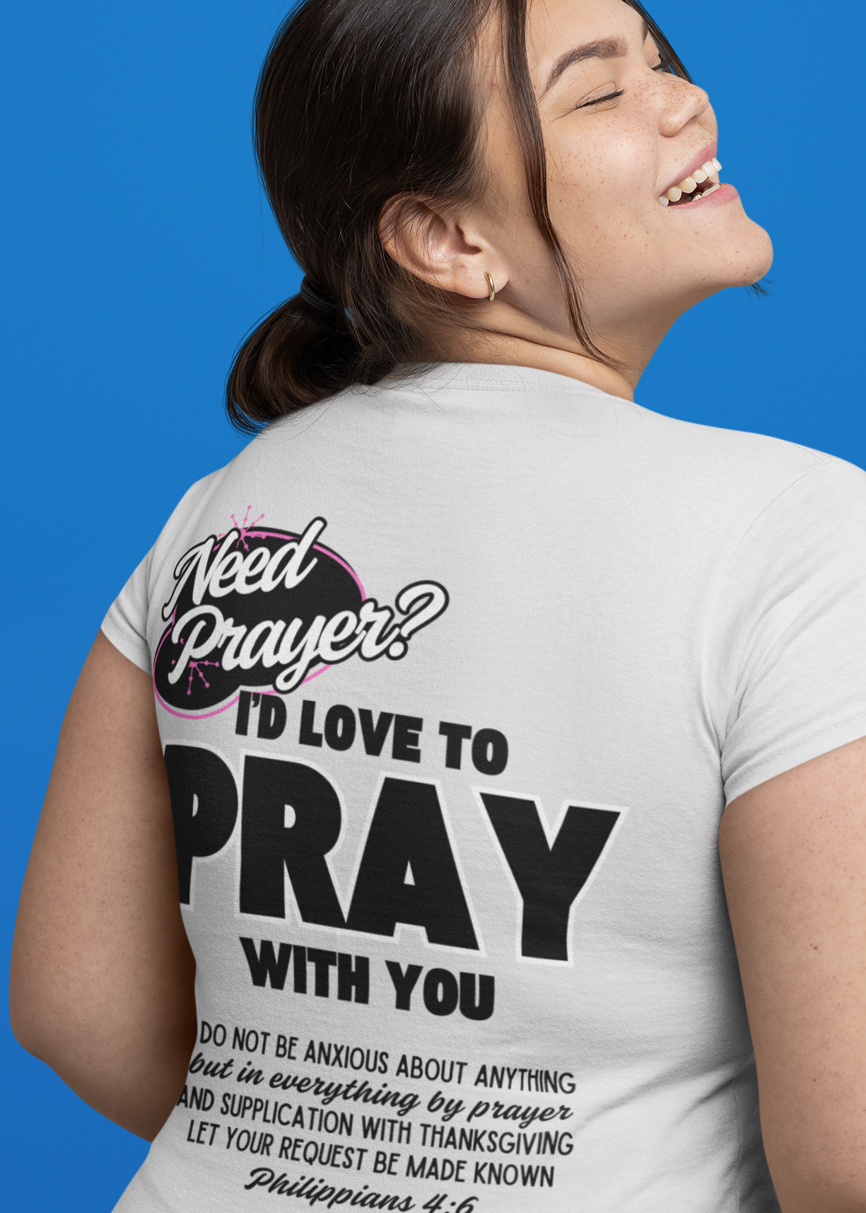 A woman shown from the back wearing a white graphic T-shirt with bold black text reading 'Need Prayer? I'D LOVE TO PRAY WITH YOU' and a Bible verse, against a solid blue background.