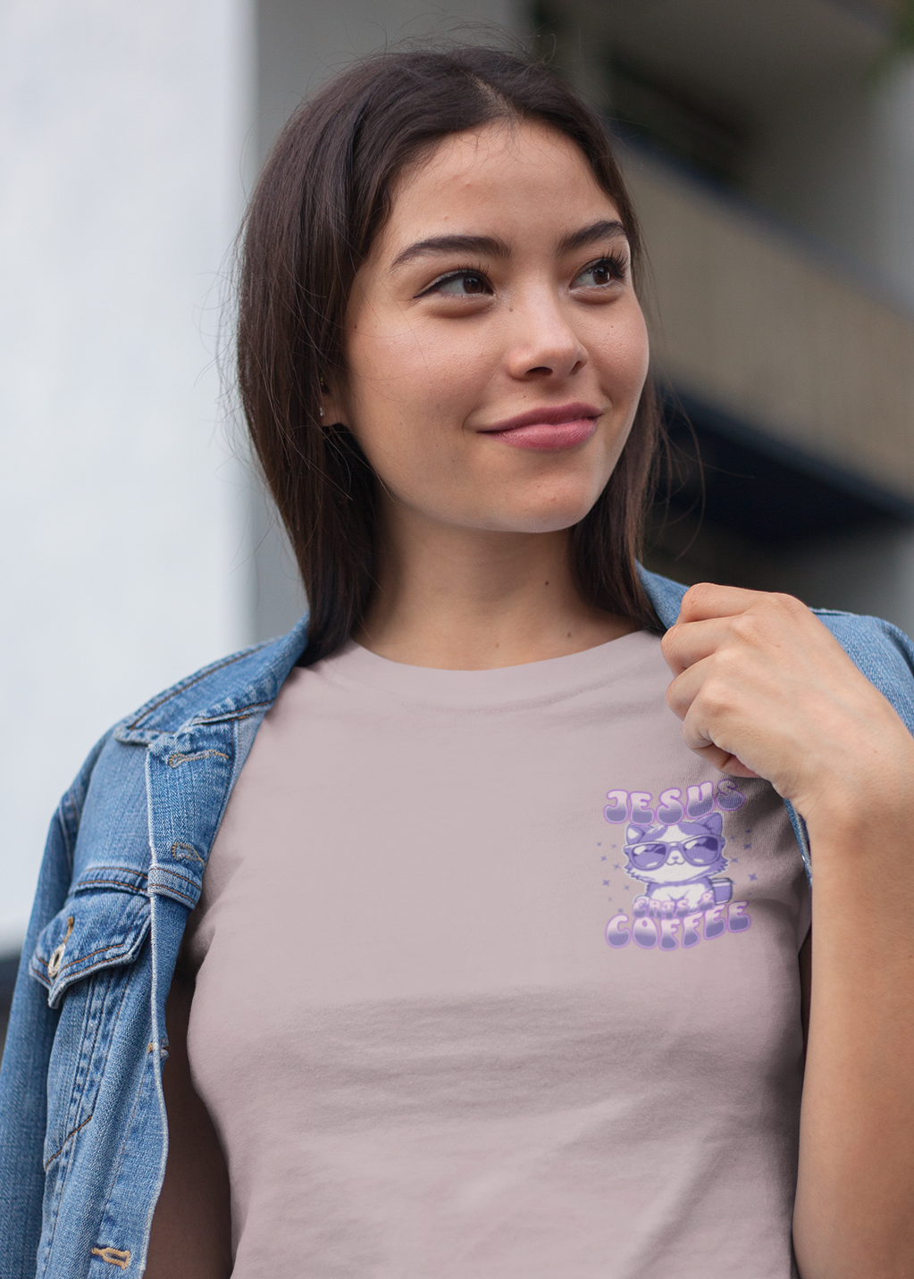 A woman wearing a light pink crewneck t-shirt with a purple cartoon graphic on the chest, styled with an open blue denim jacket, set against an outdoor urban background.