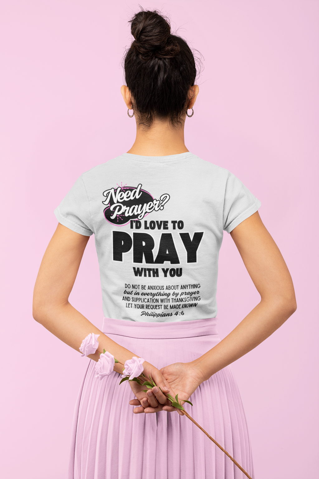 A woman shown from behind wearing a light heather gray graphic T-shirt with black text that reads 'Need Prayer? I'D Love to Pray With You'. Modely sytled in a pink pleated skirt, standing against a solid pink background.