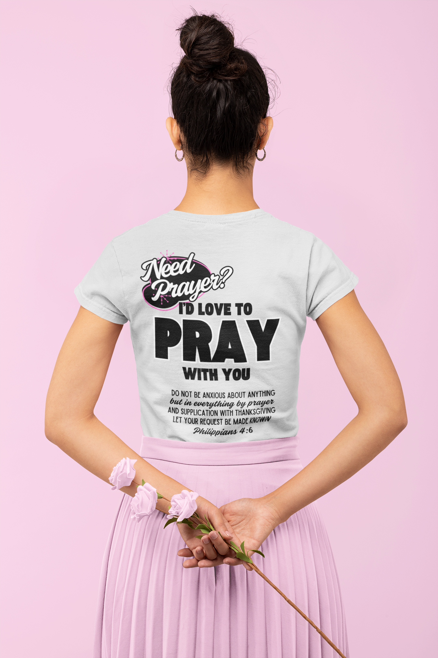 A woman shown from behind wearing a light heather gray graphic T-shirt with black text that reads 'Need Prayer? I'D Love to Pray With You'. Modely sytled in a pink pleated skirt, standing against a solid pink background.