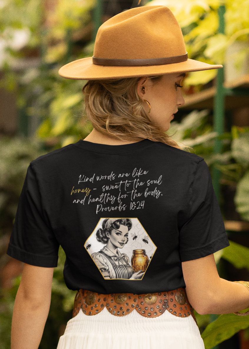 A woman viewed from behind wearing a black graphic t-shirt with white text and a white tiered maxi skirt, walking through a lush, green potted plant garden. She is wearing a tan wide-brim hat and brown flat sandals.