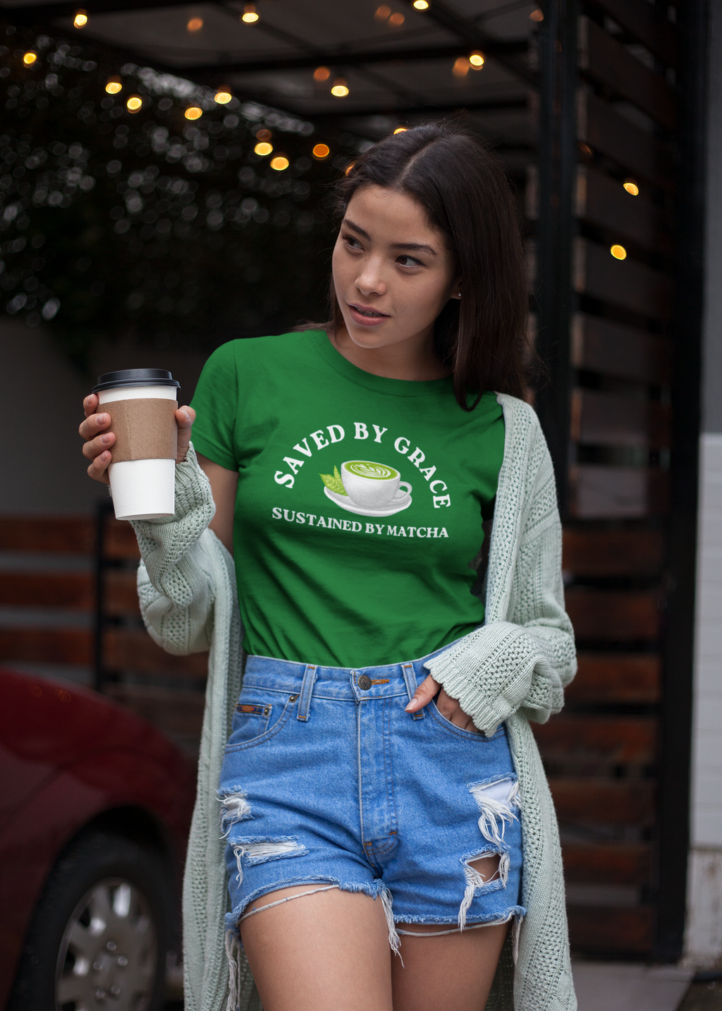 Woman wearing a kelly green t-shirt with text and graphics, holding a matcha latte outdoors. 