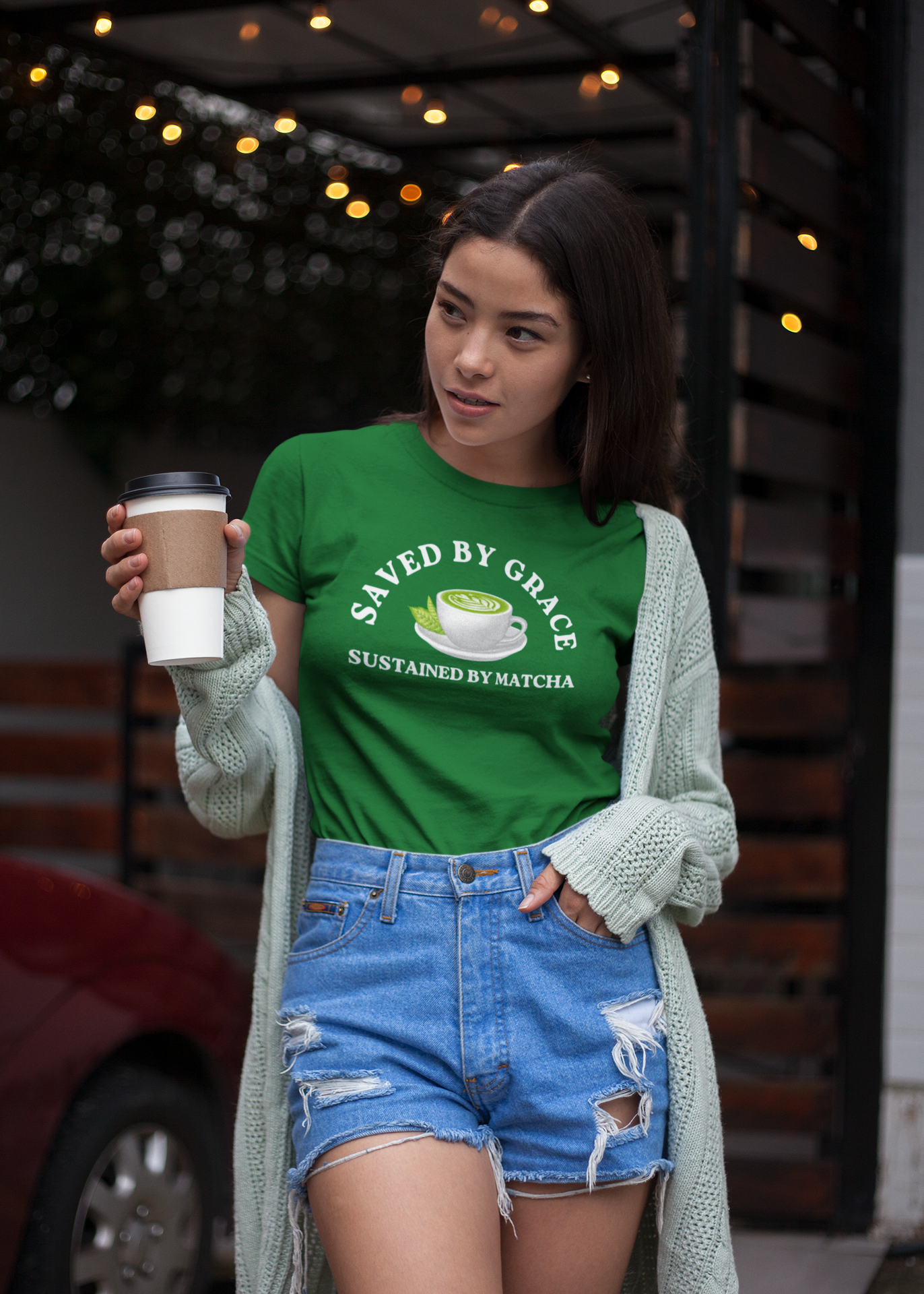Woman wearing a kelly green t-shirt with text and graphics, holding a matcha latte outdoors. 