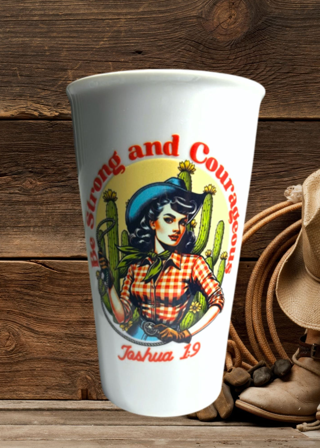 White cup with a graphic design of a woman in a cowboy hat and plaid shirt, surrounded by cacti, on a wooden surface with cowboy boots and hat.