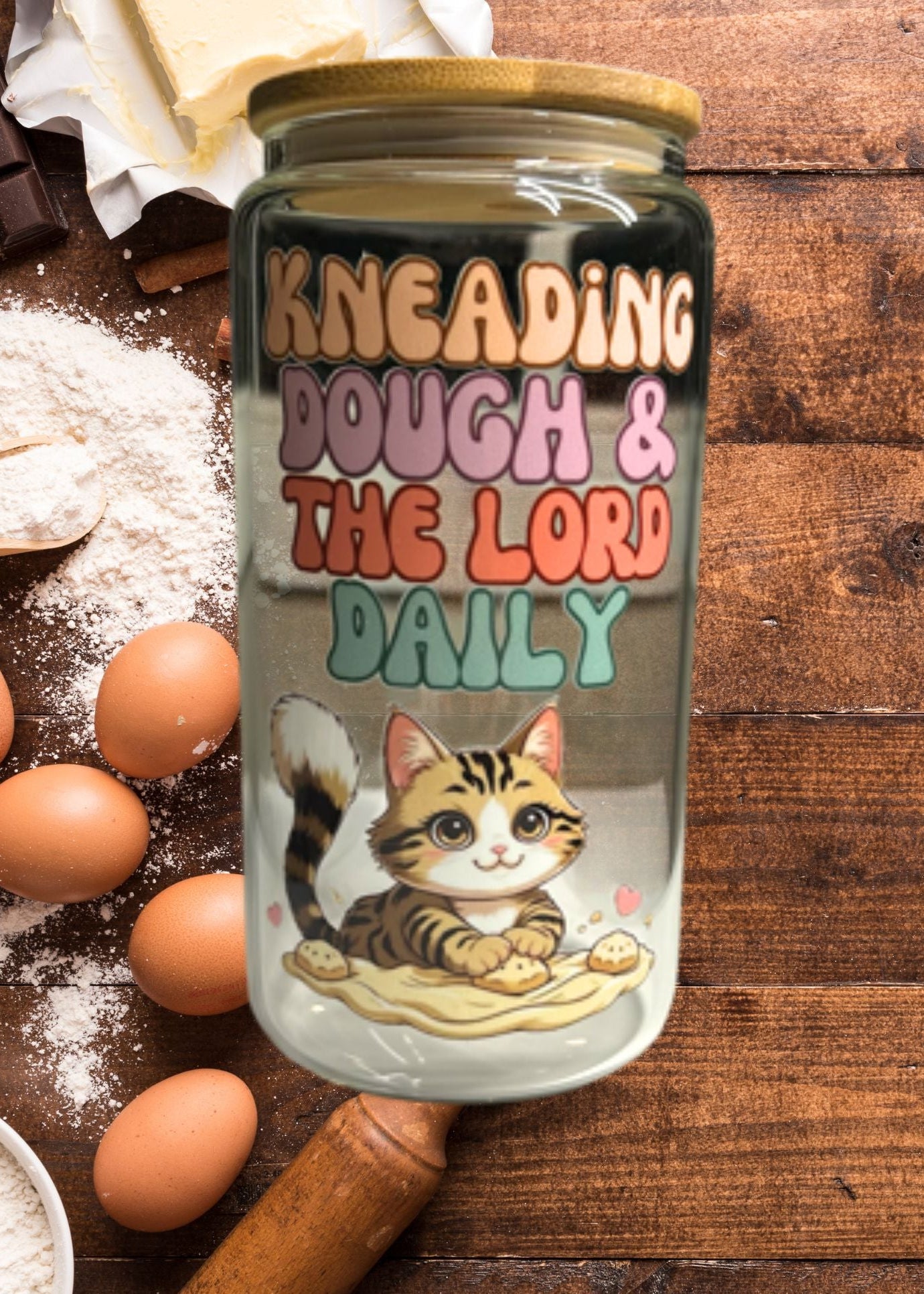 Jar with a cat illustration and text 'Kneading Dough & The Lord Daily' on a wooden surface with flour and eggs.