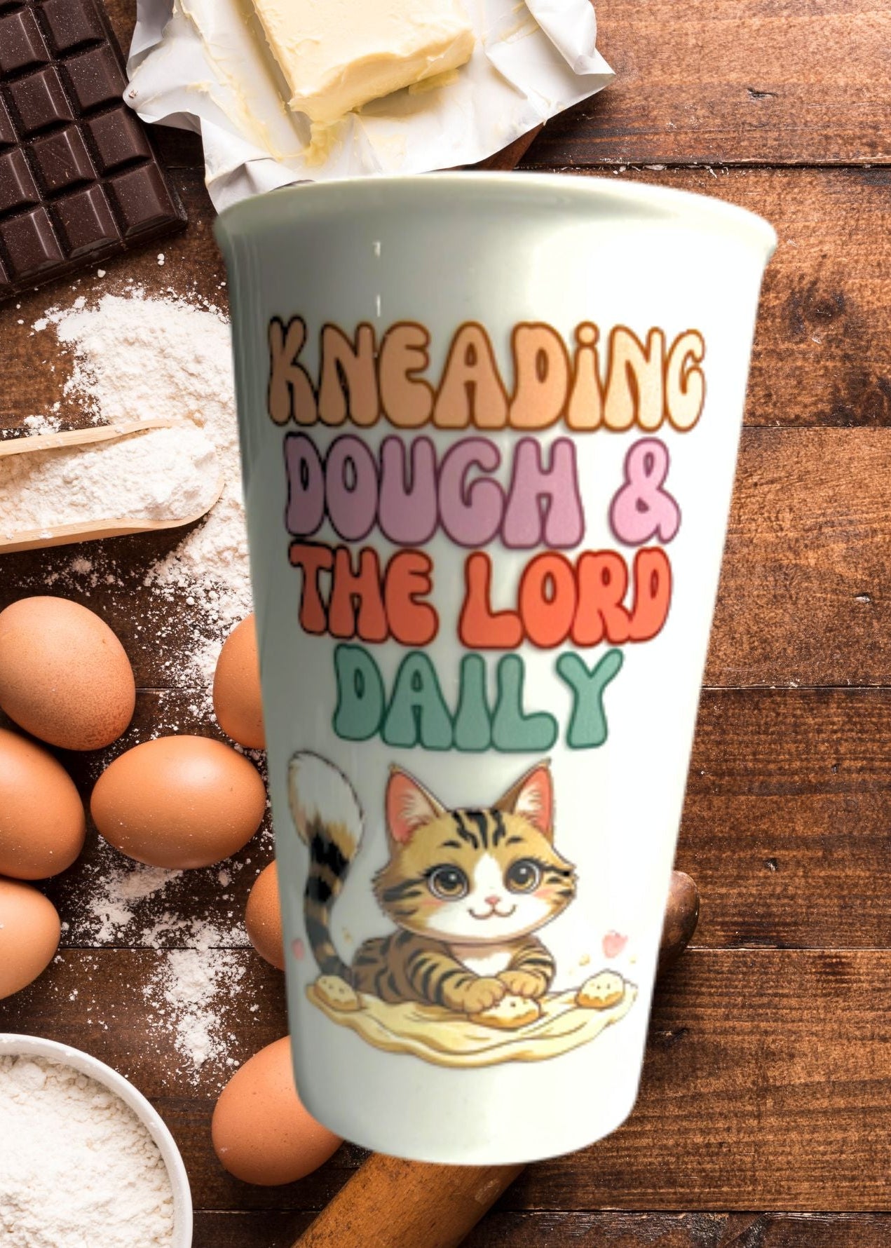 Cup with a cat illustration and text 'Kneading Dough & The Lord Daily' on a wooden surface with baking ingredients.