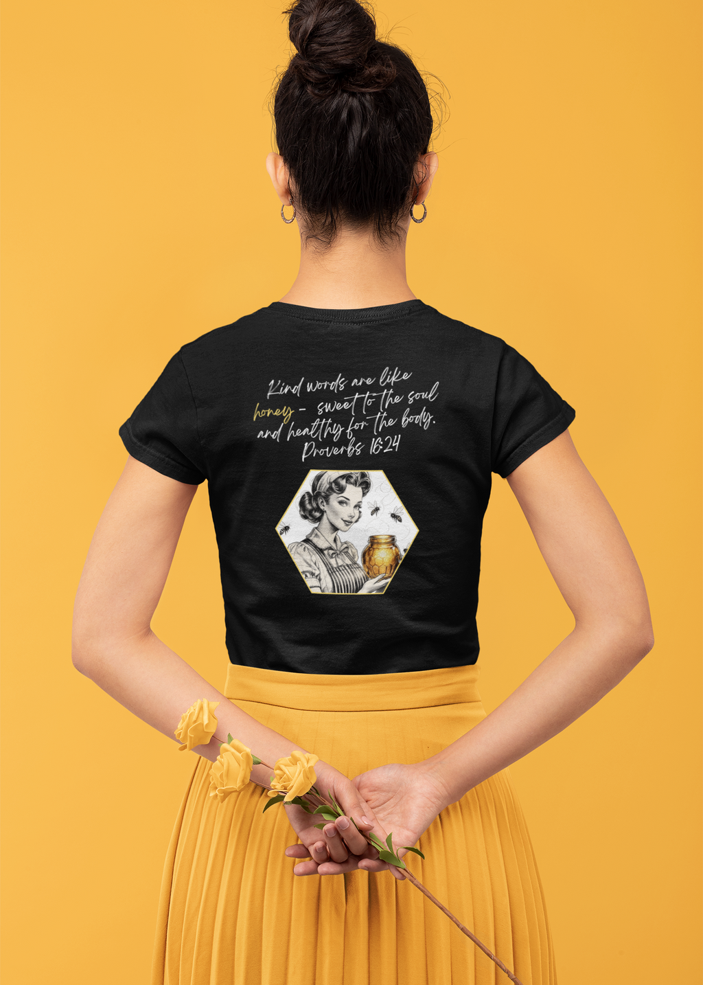 Black Christian graphic tee with retro woman holding honey jar, bees flying around, and Proverbs 16:24 scripture about kind words. Woman is wearing the shirt holding yellow roses and wearing a yellow skirt. Messy bun. 