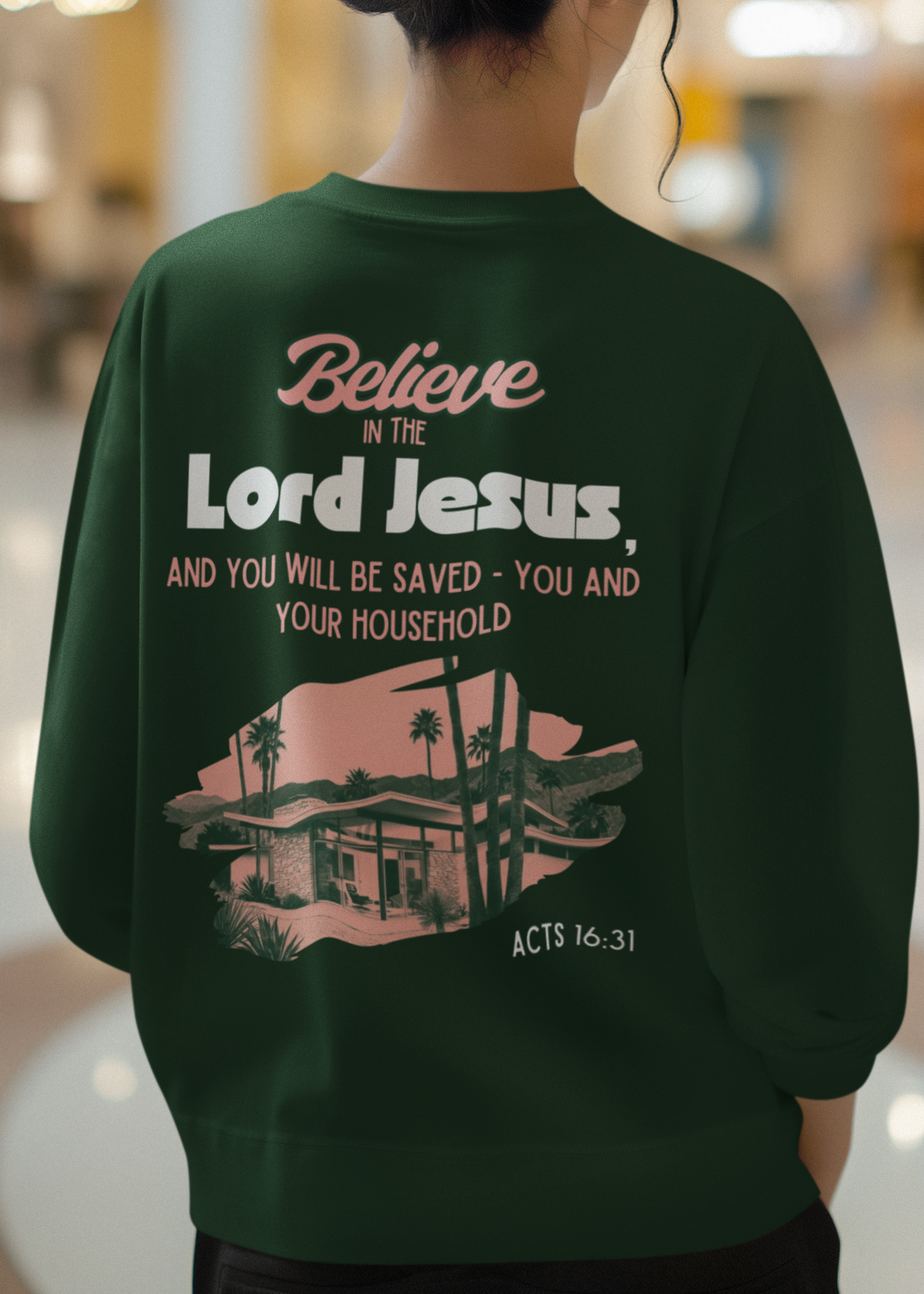 A person viewed from behind wearing a dark green sweatshirt with a large back graphic that reads, 'Believe in the Lord Jesus, and you will be saved - you and your household' above a pink and green house illustration and the text 'ACTS 16:31'. The setting appears to be a blurred indoor mall or public space.