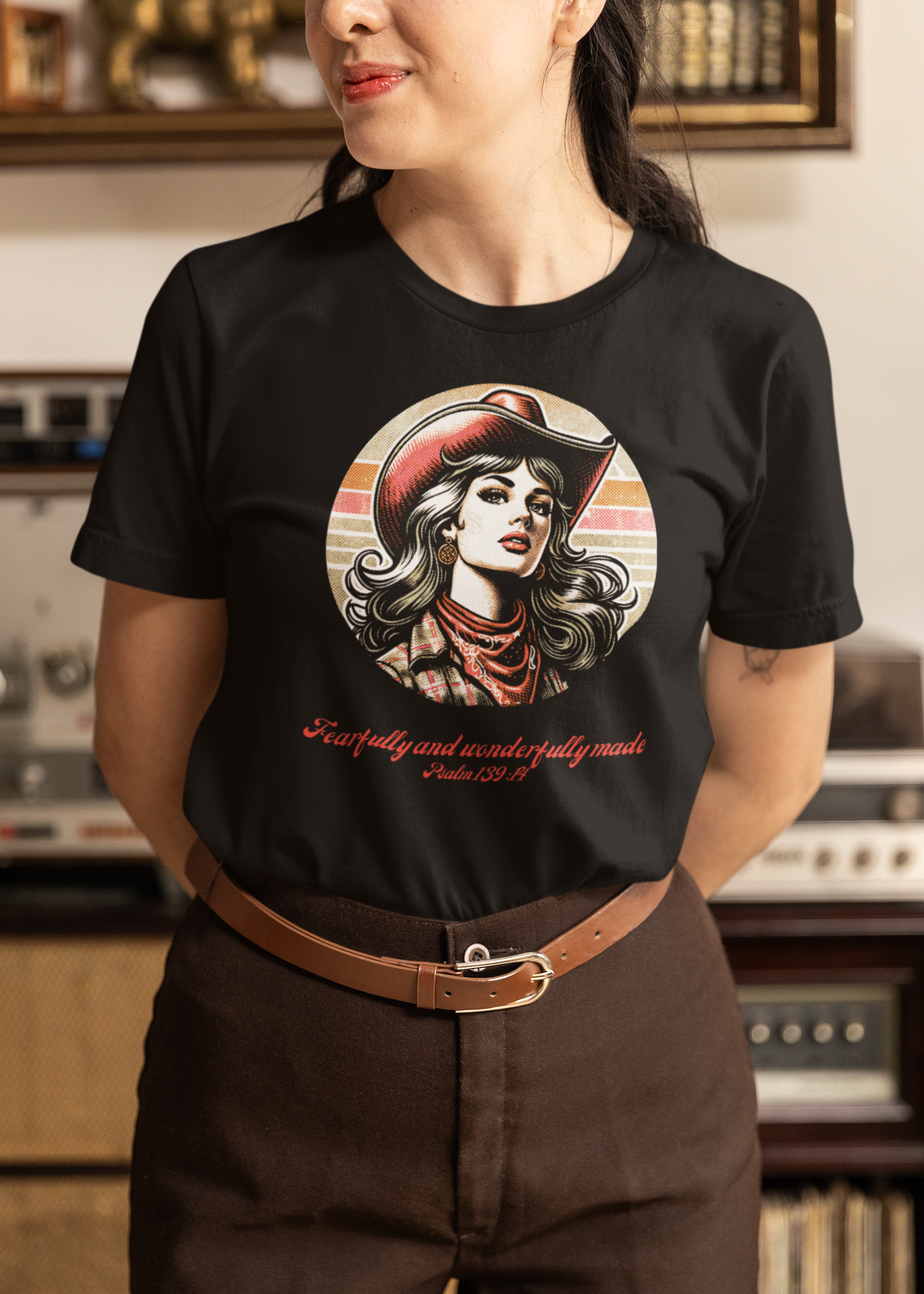 Woman wearing black 70s-style cowgirl t-shirt with Psalm 139:14 design, paired with brown pants, sitting in retro living room with record player, face cropped