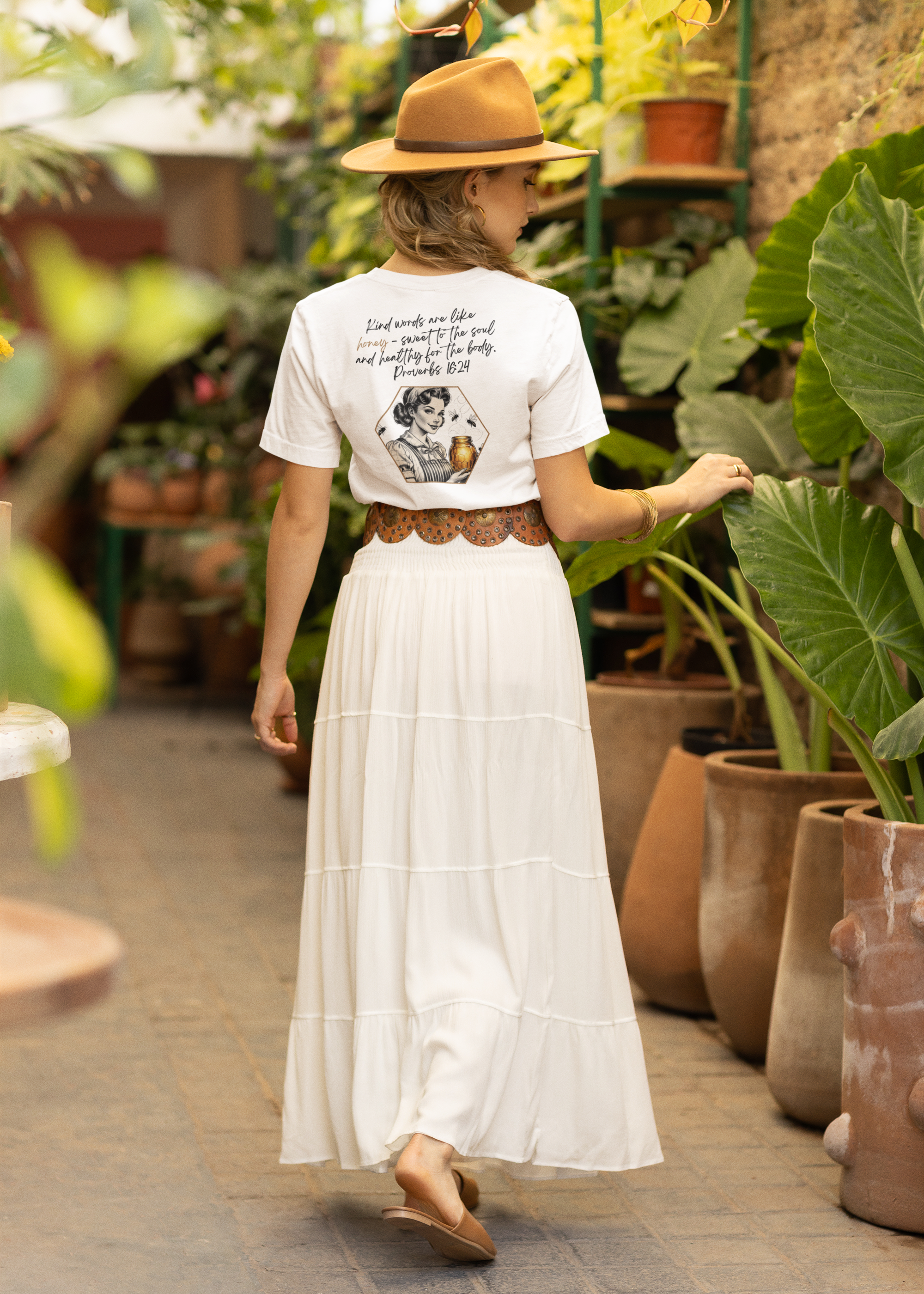 Woman in a white dress and hat walking through a greenhouse filled with plants.
