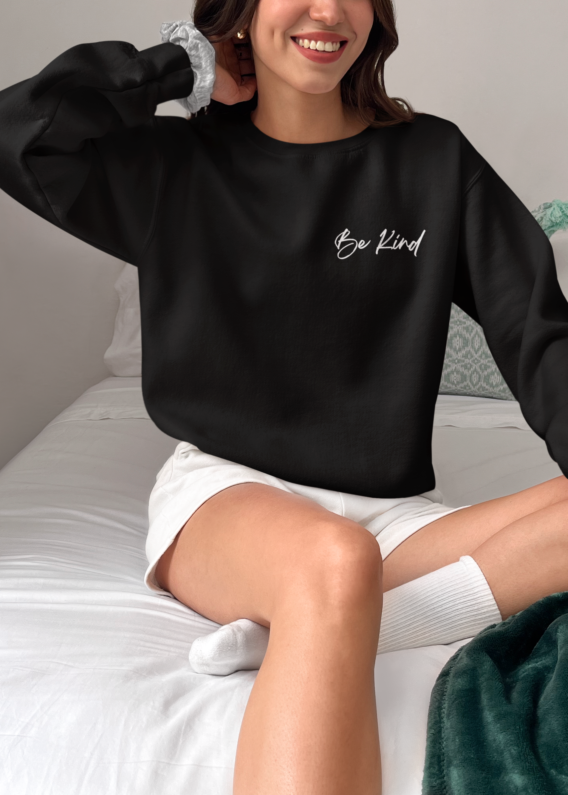 A woman sitting on a bed wearing a black crewneck sweatshirt with 'Be Kind' written in white script on the chest, paired with white shorts and white socks.