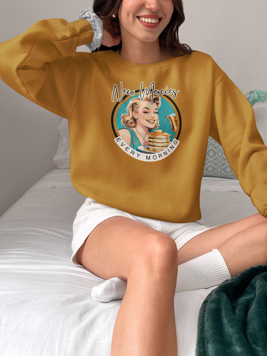 A woman sitting on a bed wearing a mustard yellow sweatshirt with a retro graphic of a woman holding a stack of pancakes and the text 'New Meowicz Every Morning'.