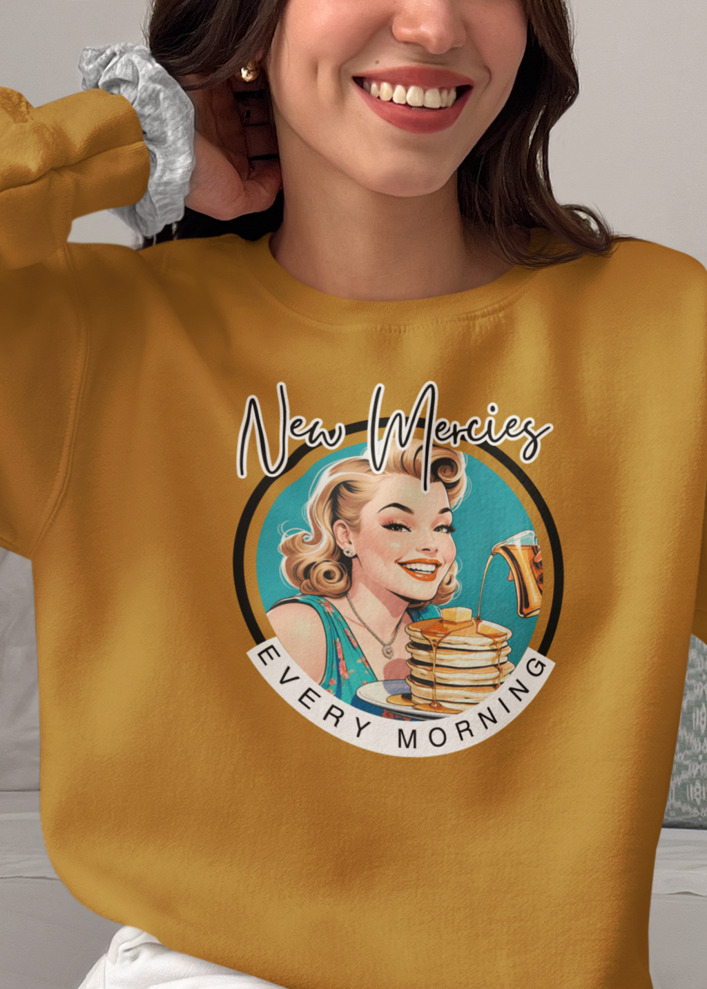 A woman sitting on a bed wearing a mustard yellow sweatshirt with a retro graphic of a woman holding a stack of pancakes and the text 'New Meowicz Every Morning'.