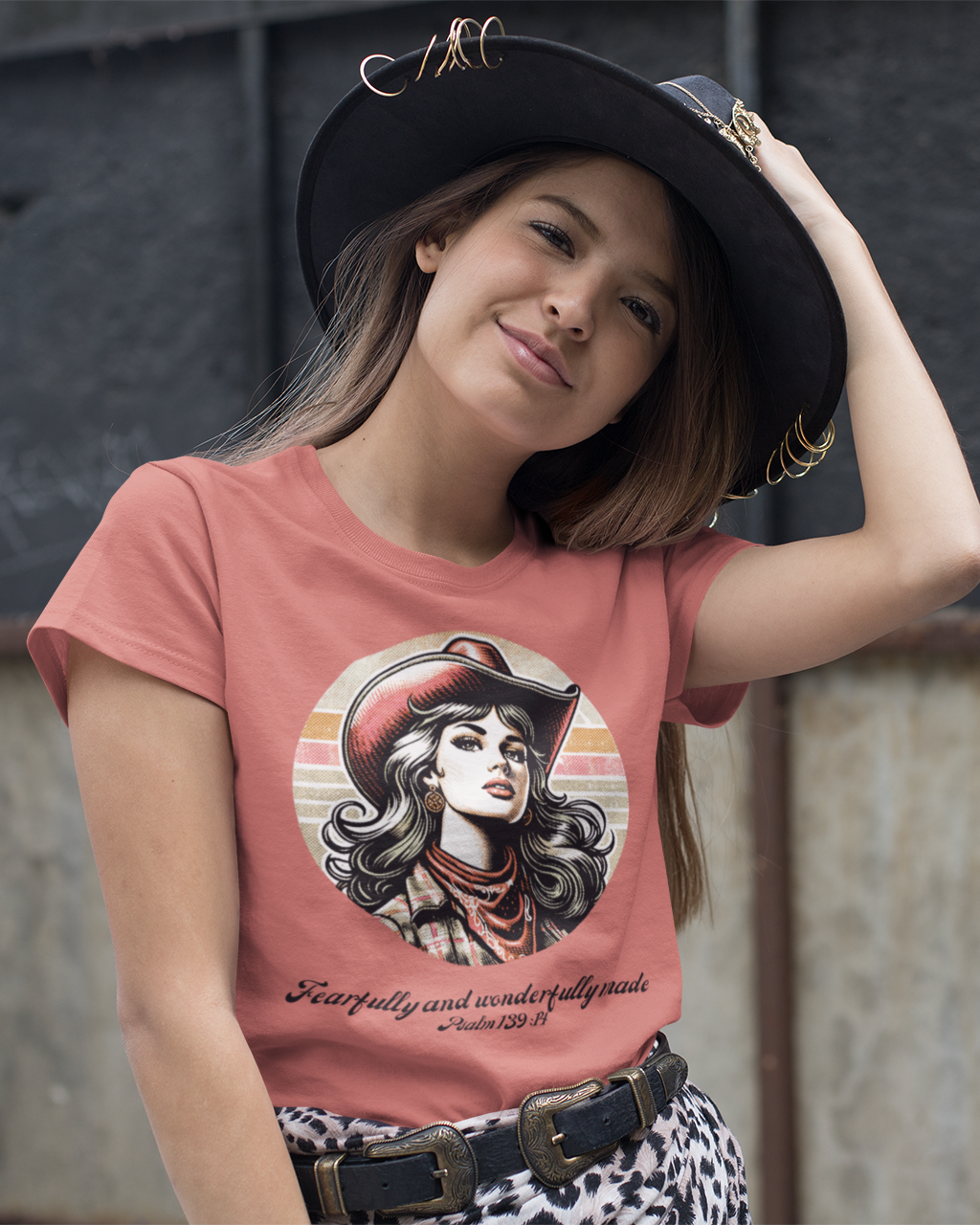Woman wearing a pink t-shirt with a graphic design of a woman in a cowboy hat, standing against a textured wall.