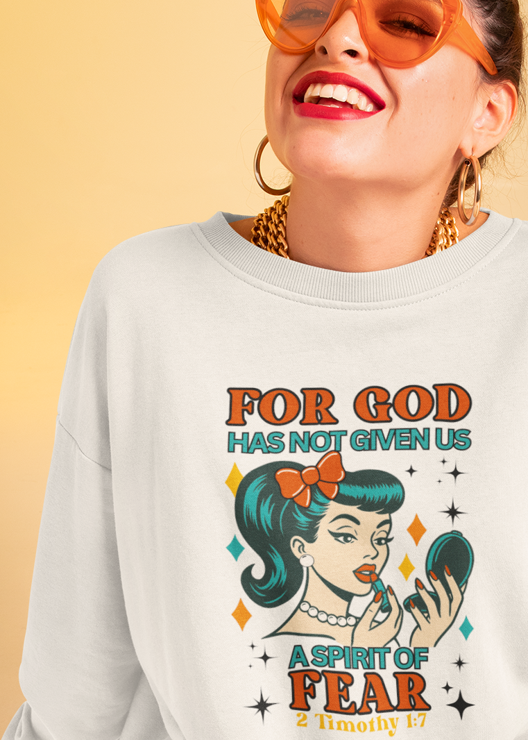 A woman wearing a white graphic sweatshirt with a retro-style illustration and the text 'FOR GOD HAS NOT GIVEN US A SPIRIT OF FEAR', paired with tan pants and orange-tinted sunglasses, posed against a warm beige background.