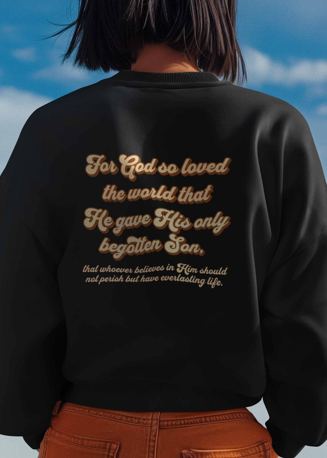 A person viewed from behind wearing a black crewneck sweatshirt with gold cursive text on the back, paired with orange-brown pants, set against a blue sky background.