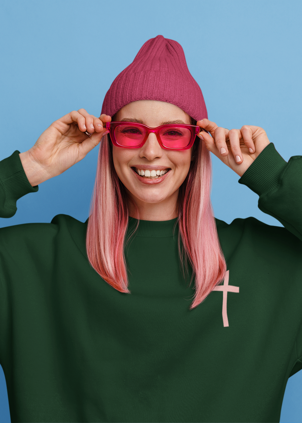 A smiling woman with pink hair wears a dark green sweatshirt, a magenta beanie, and pink sunglasses against a solid blue background.