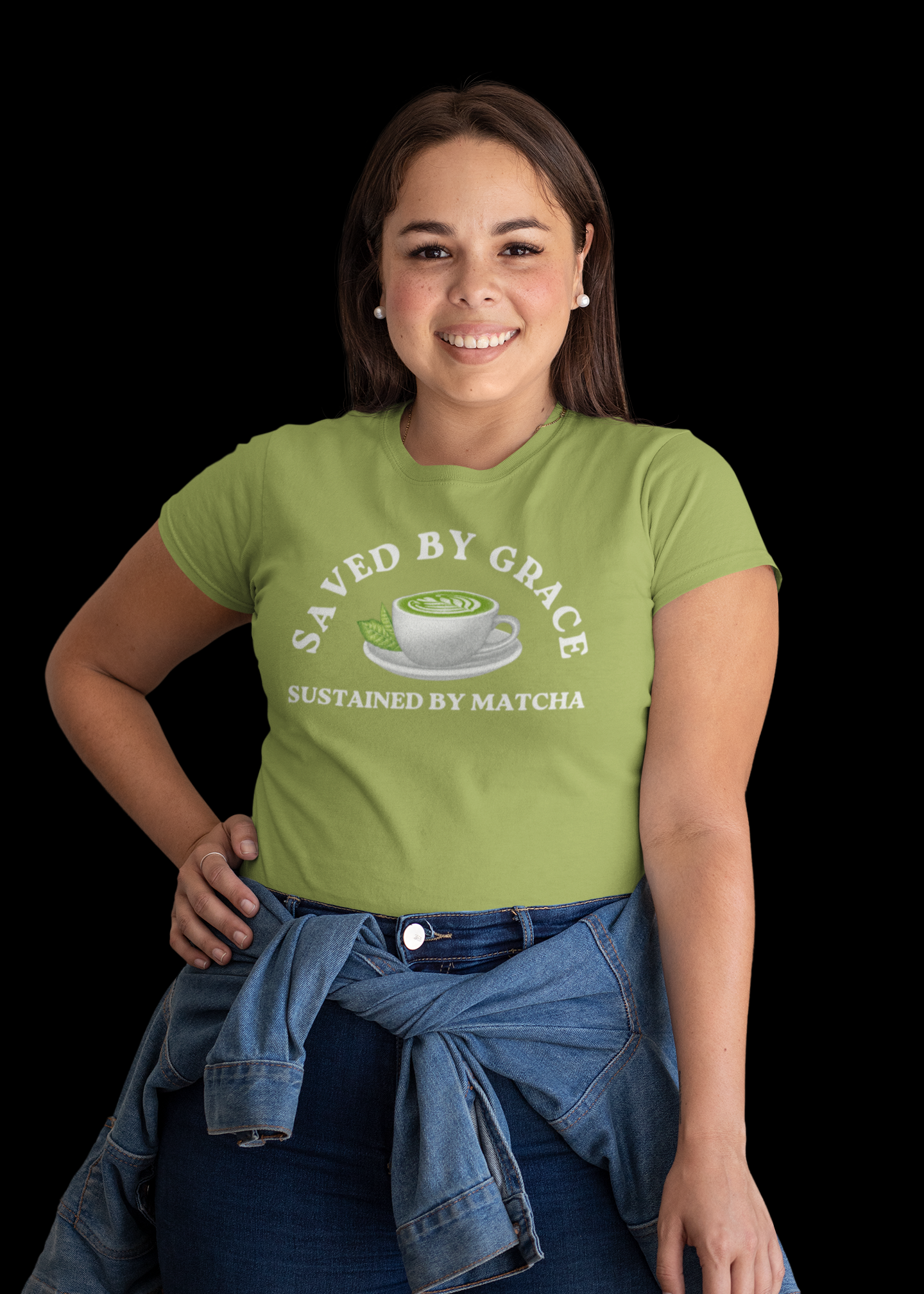 A woman modeling a light green graphic t-shirt with a white print that reads 'SAVED BY GRACE SUSTAINED BY MATCHA' with a matcha cup illustration. She has a blue denim jacket tied around her waist and is posed against a solid black background.